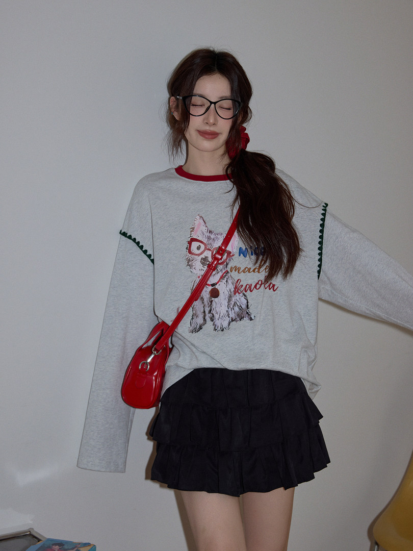 Model wearing a gray graphic long-sleeve top with a red crossbody bag and black pleated skirt