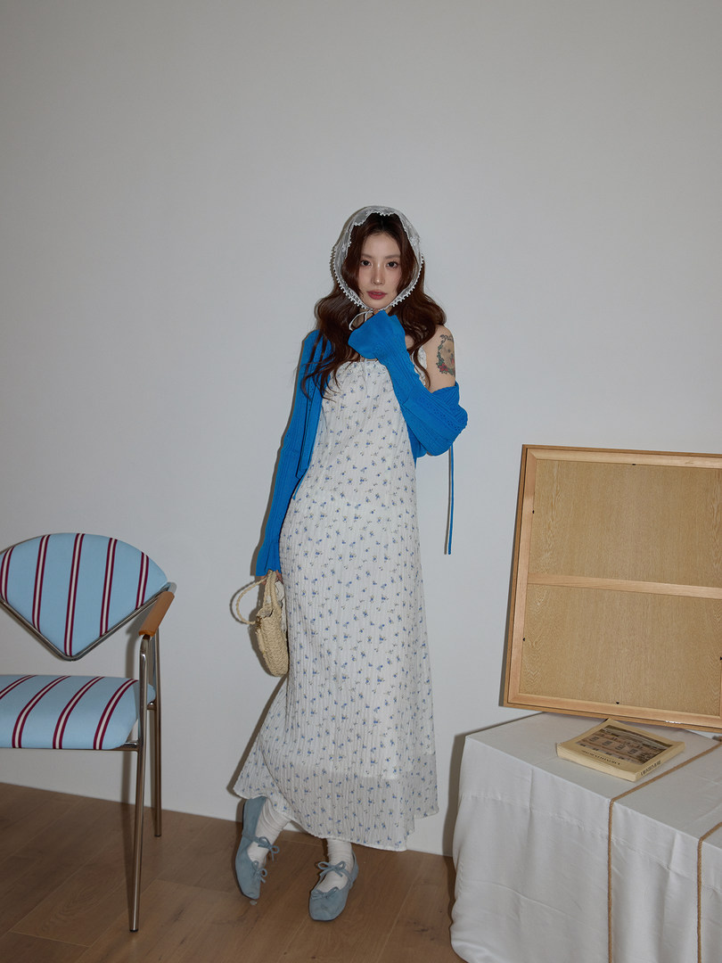Model wearing a long floral dress with a bright blue cardigan, bonnet, and woven bag