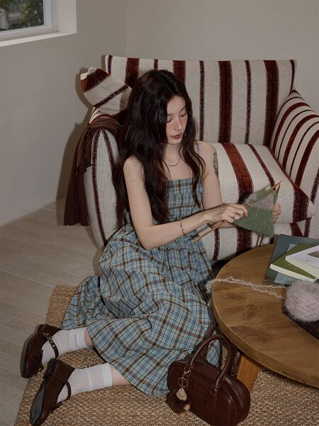 Woman wearing a plaid sleeveless dress while sitting indoors with a brown handbag and shoes