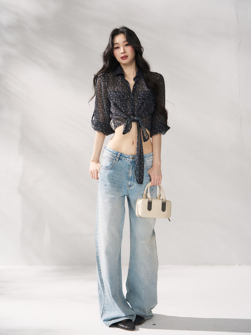 Model wearing a sheer black tie-front blouse with light wash wide-leg jeans and a small beige handbag