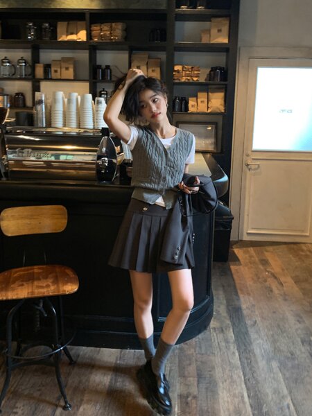 Model wearing a gray knit vest over a white T-shirt with a black pleated skirt and loafers