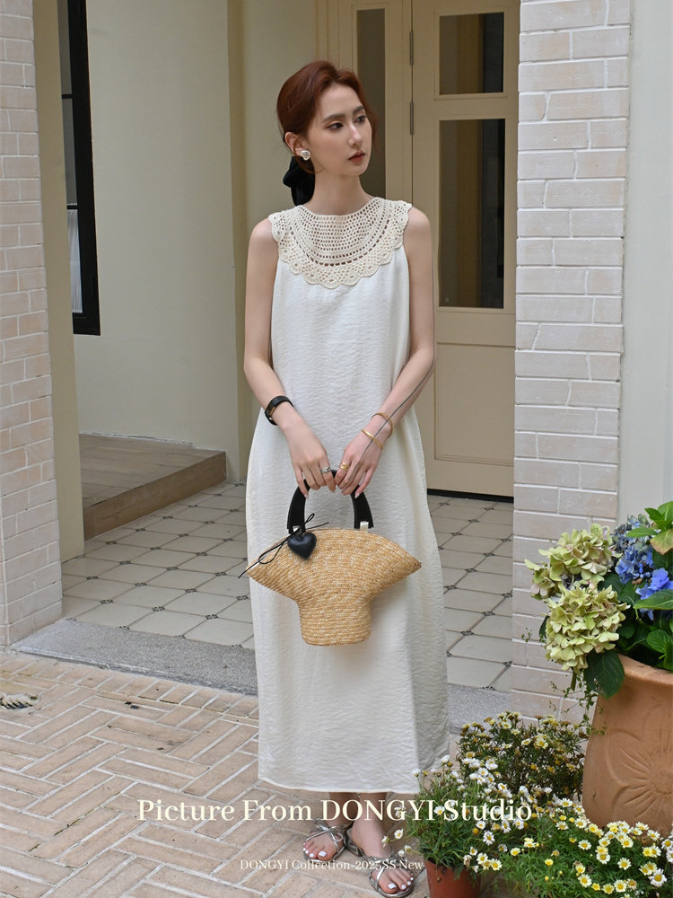 Model wearing a long white sleeveless dress with a lace collar while holding a woven handbag