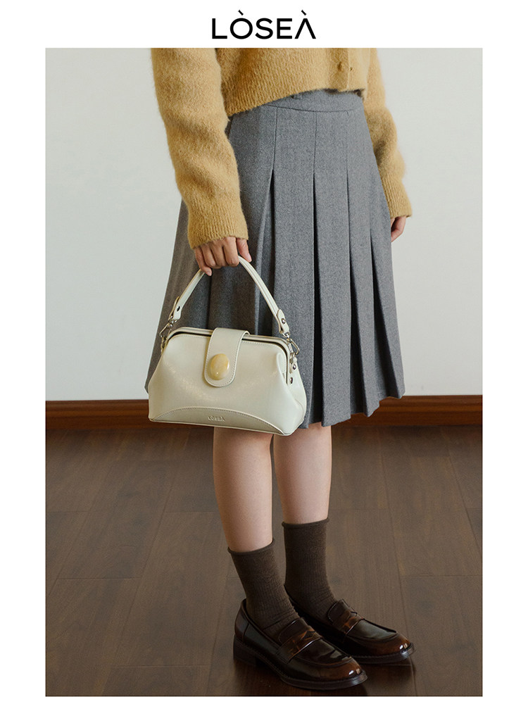 Model wearing a gray pleated skirt and holding a cream handbag