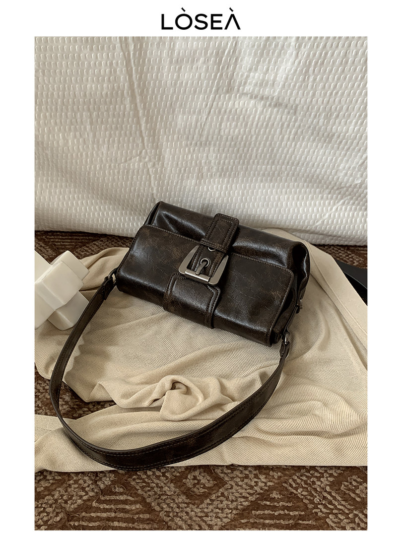Dark brown leather shoulder bag with buckle detail on a neutral fabric backdrop
