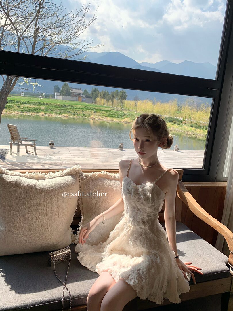 Model wearing a cream ruffled spaghetti-strap dress sitting indoors by a large window
