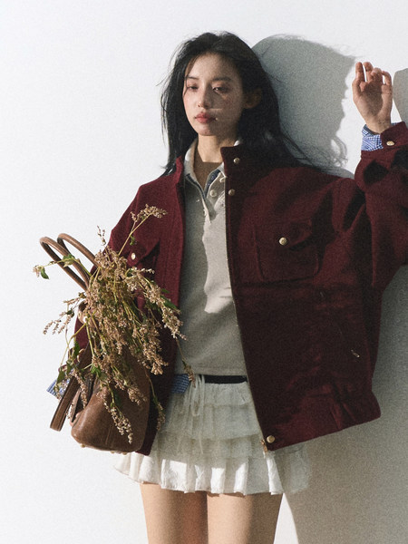 Model wearing a burgundy jacket over a light layered skirt, holding a brown handbag