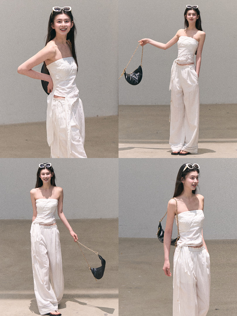 Model wearing a white strapless top and loose white wide-leg pants with a black chain bag