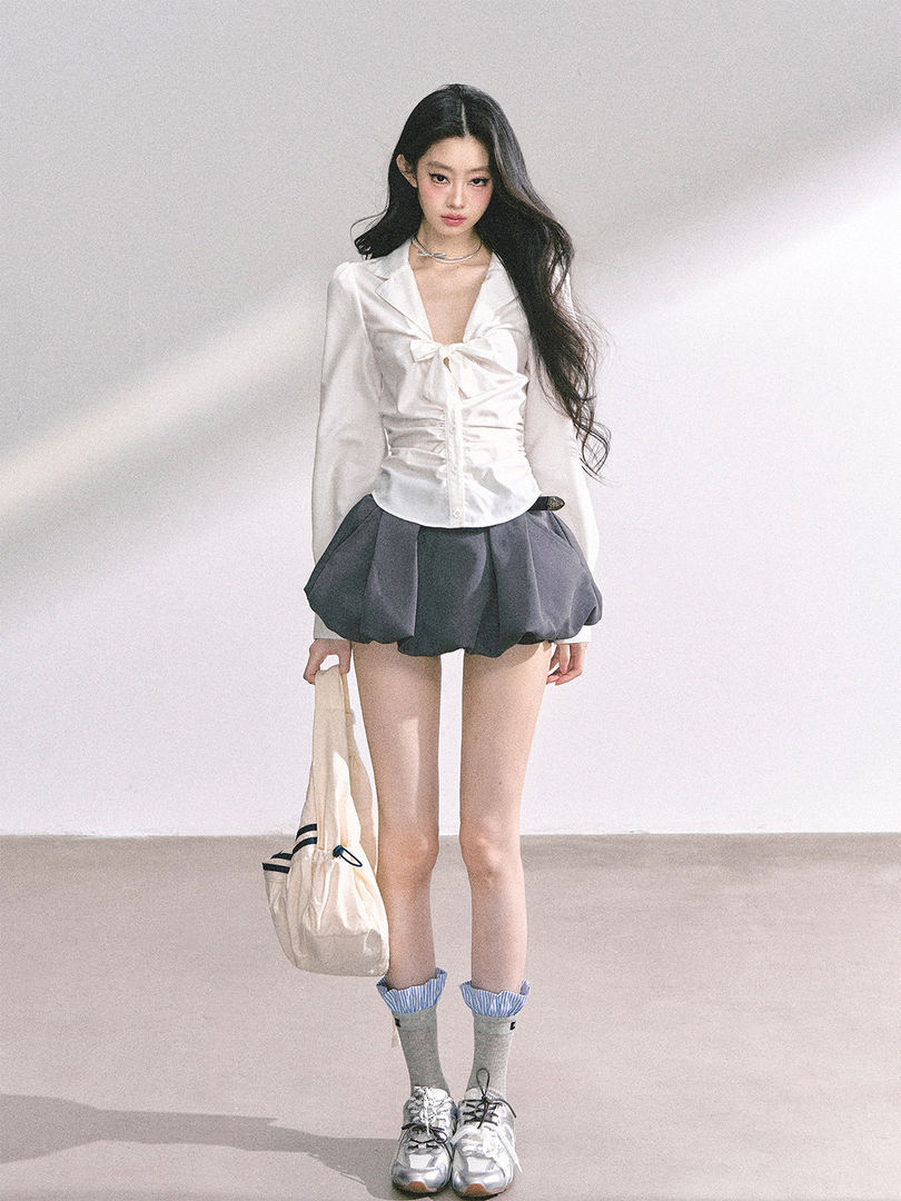 Model wearing a white tied blouse with a dark bubble skirt and silver sneakers.