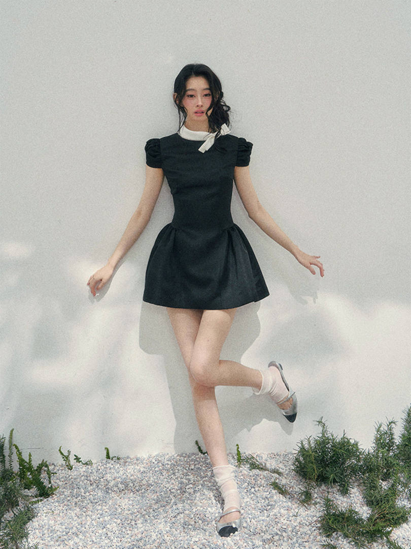 Model wearing a black short dress with puff sleeves and a white neck tie