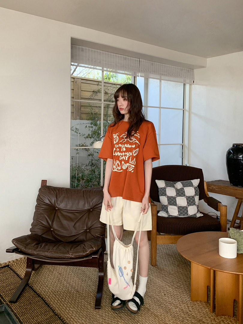 Model wearing an orange graphic T-shirt with cream shorts and a drawstring bag