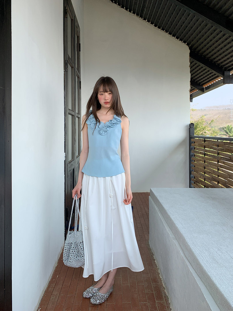 Model wearing a light blue ruffled sleeveless top with a white midi skirt and woven handbag