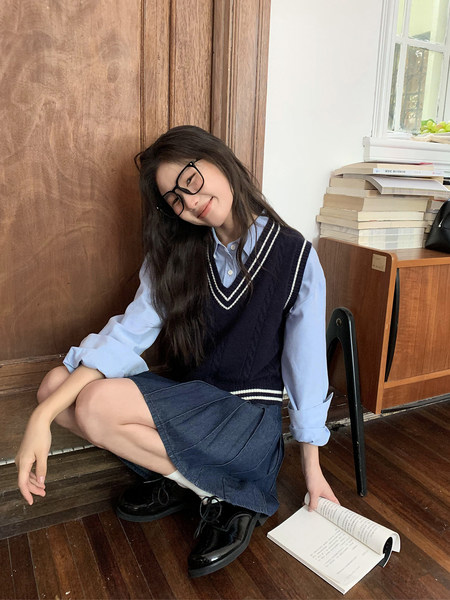 Young woman wearing a navy sweater vest over a light blue shirt with a denim skirt and black shoes