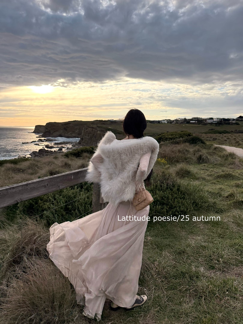 Woman wearing a flowing blush dress with a white faux fur wrap on a coastal cliff at sunset