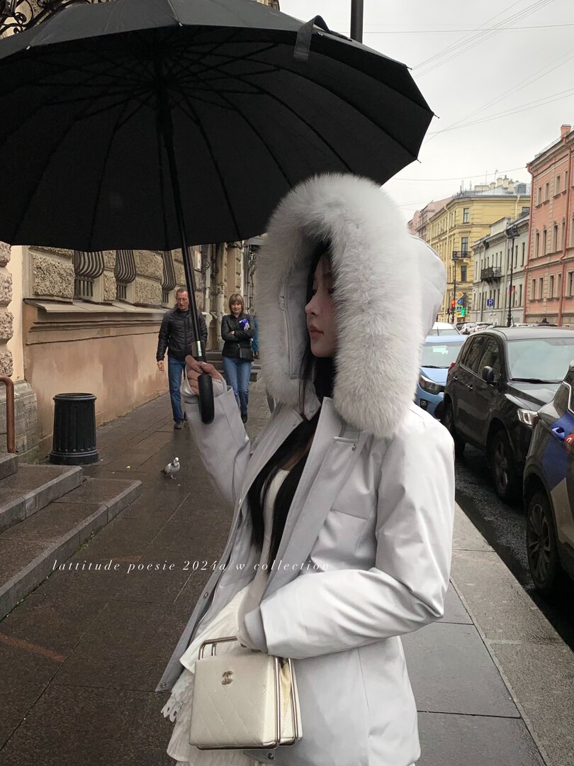 Woman wearing a white fur-trim hooded coat and carrying a cream handbag under a black umbrella