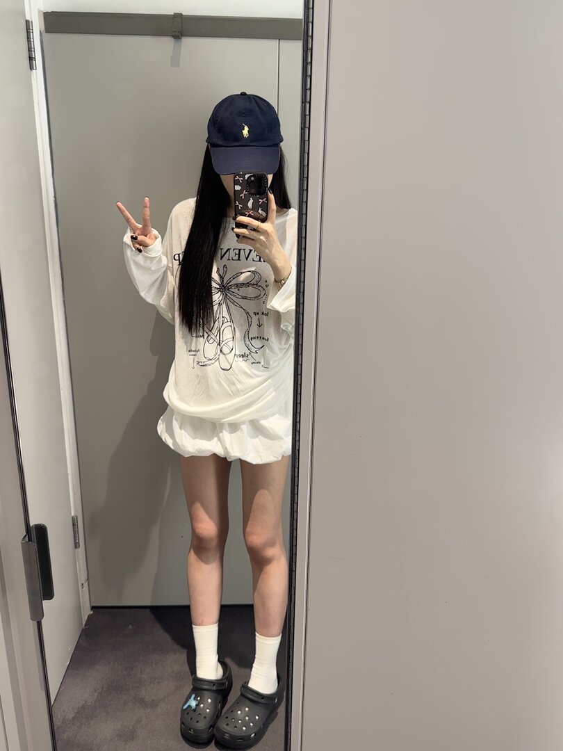 Mirror selfie of a model wearing a white graphic long-sleeve top, white shorts, navy cap, and black Crocs