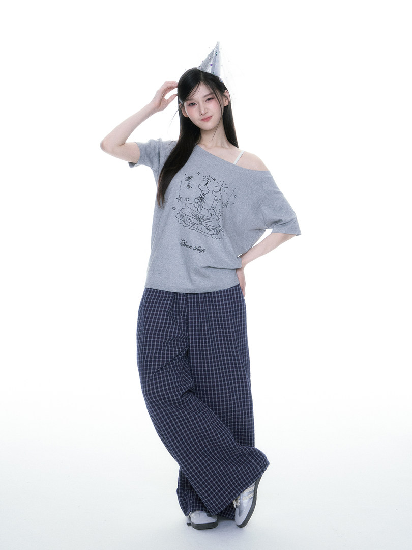 Model wearing a gray graphic off-shoulder top with plaid lounge pants and a party hat
