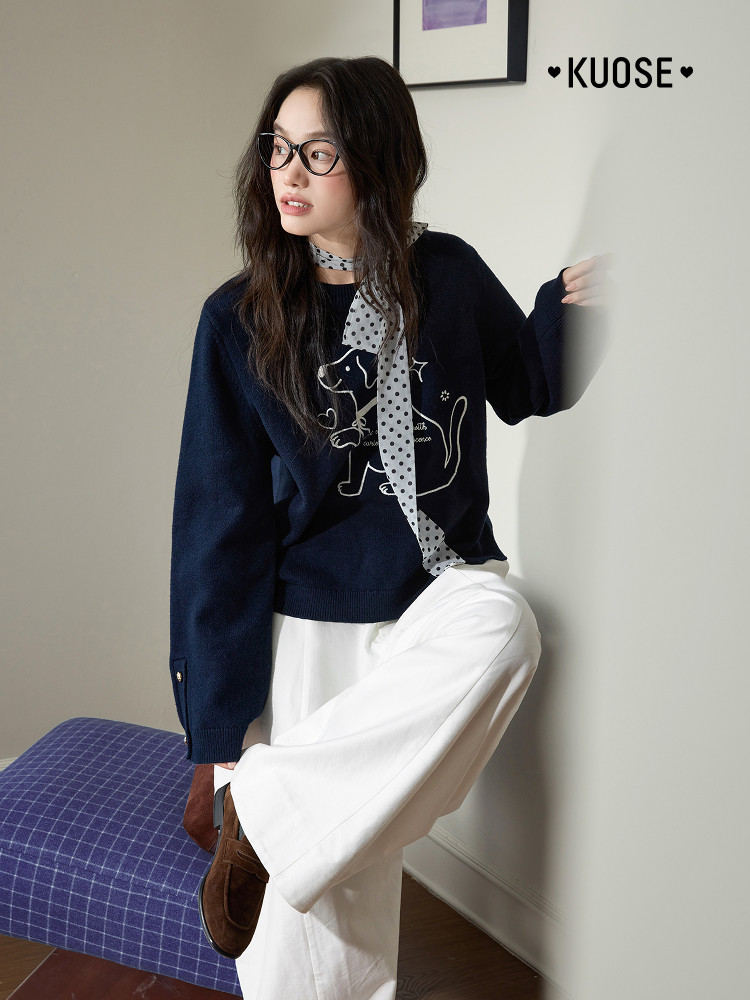 Model wearing a navy graphic sweater with a polka dot scarf, white wide-leg pants, and brown loafers