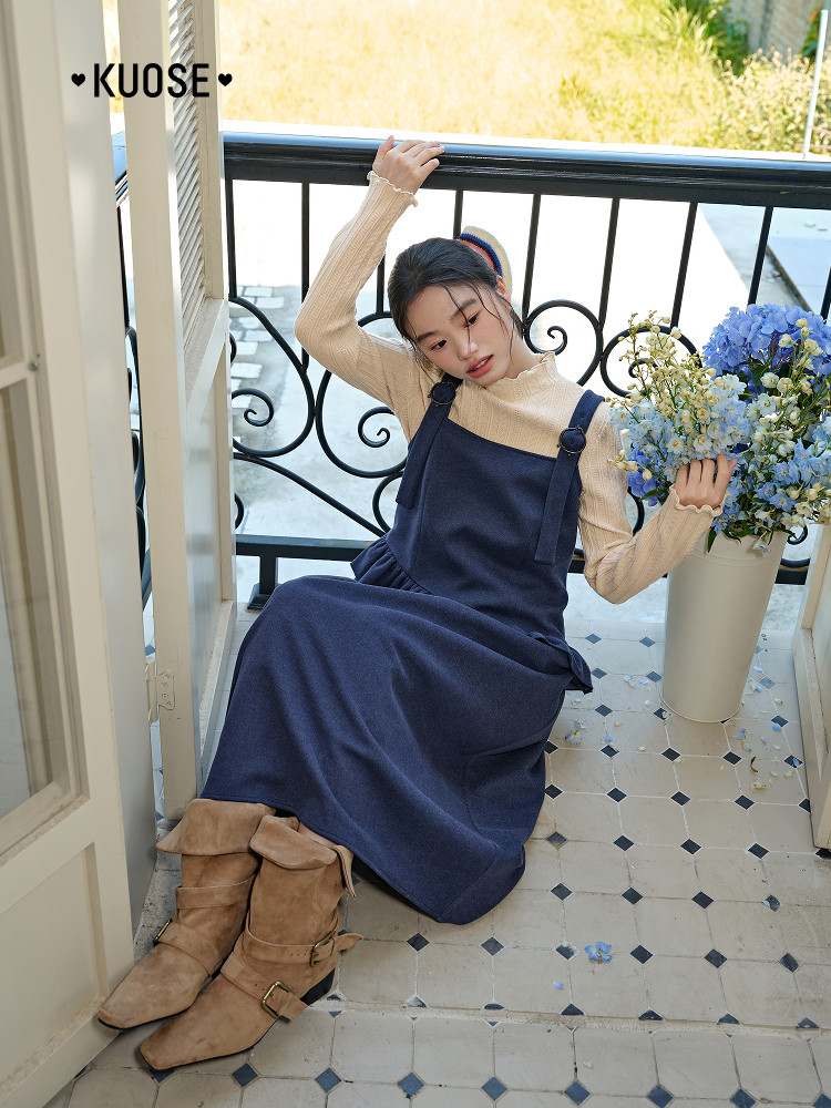 Model wearing a navy pinafore dress over a beige long-sleeve top with tan boots