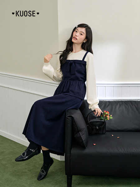 Model wearing a navy pinafore dress over a cream long-sleeve top with black loafers