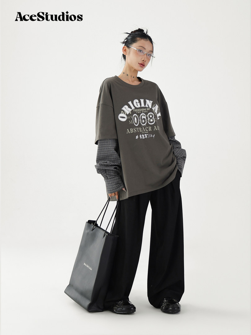 Model wearing an oversized graphic T-shirt with layered check sleeves, wide black pants, and a tote bag