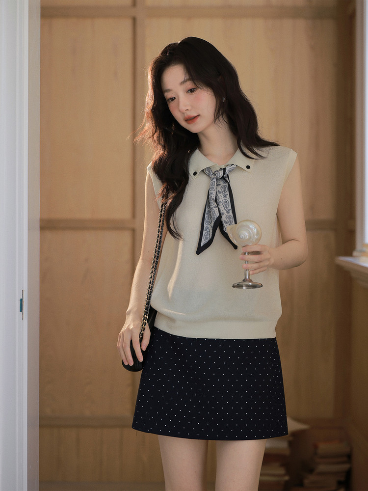Model wearing a cream sleeveless knit top with a neck scarf and a navy polka dot mini skirt