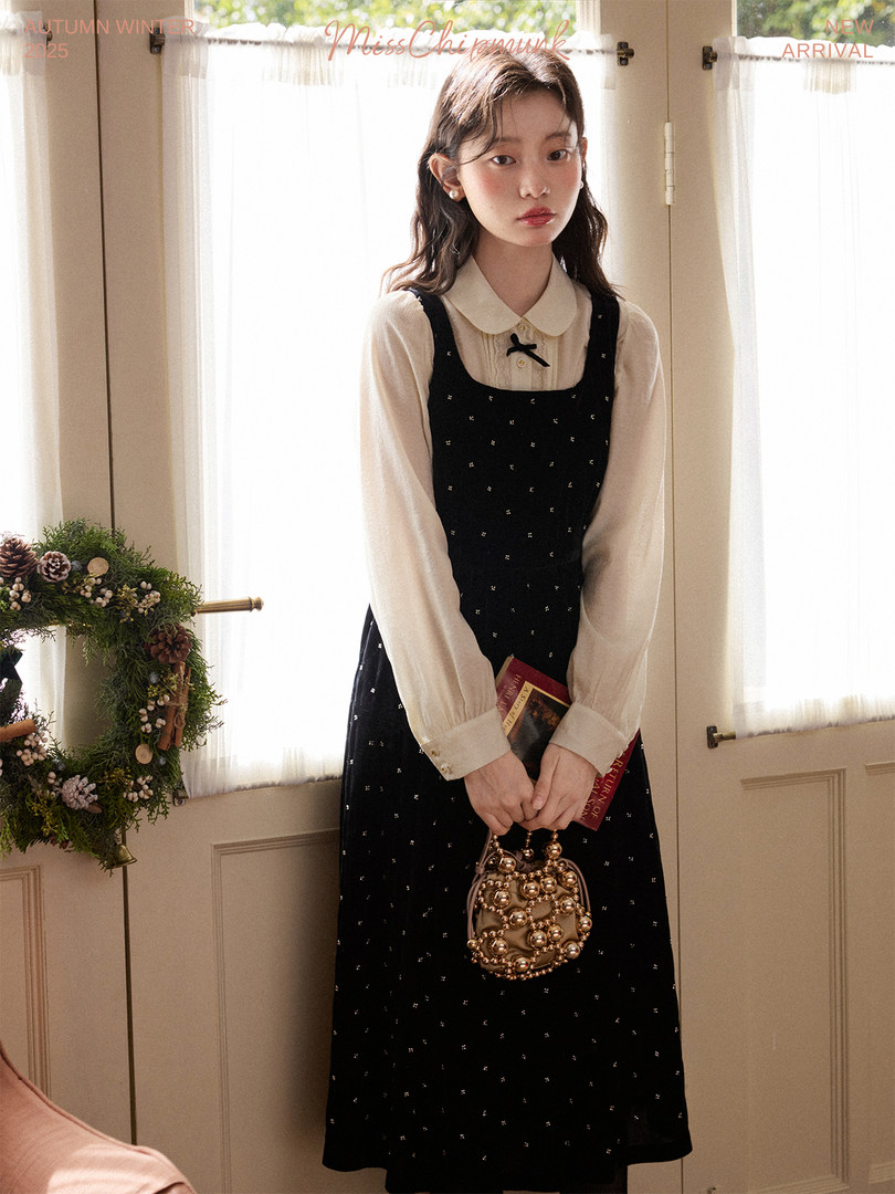 Model wearing a black pinafore dress over a cream blouse with a small handbag