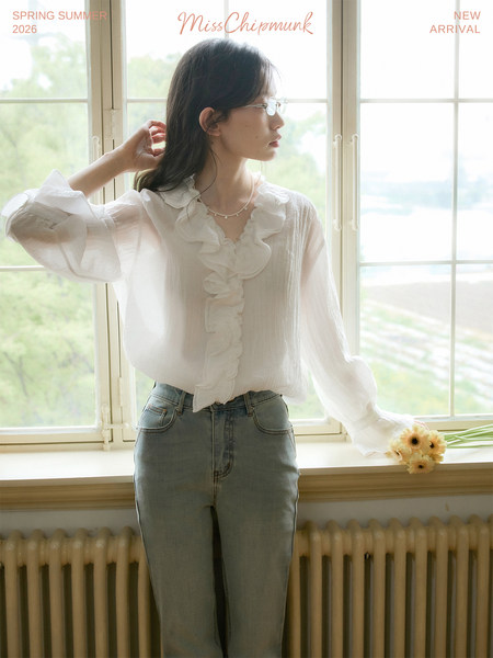 Model wearing a white ruffled sheer blouse with light blue jeans indoors by a window