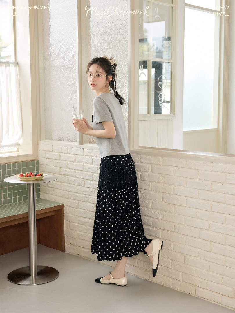 Model wearing a gray T-shirt with a black polka dot skirt and flats in a bright cafe setting