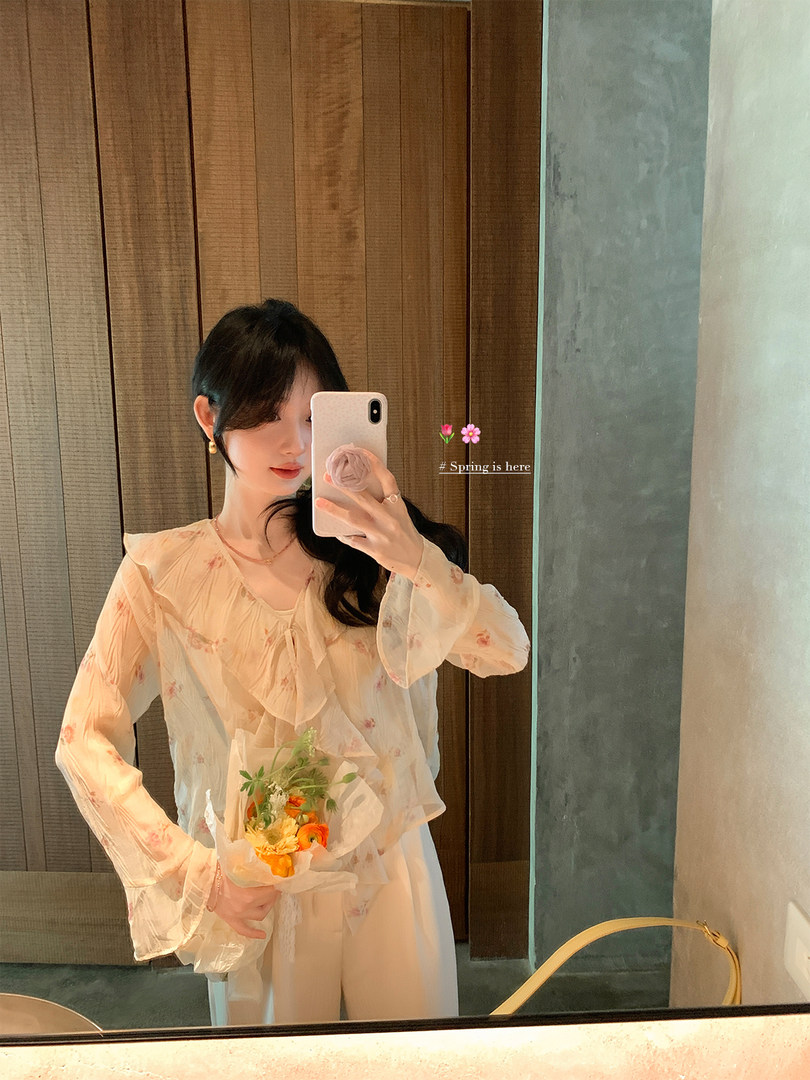 Woman wearing a sheer floral blouse and light pants in a mirror selfie