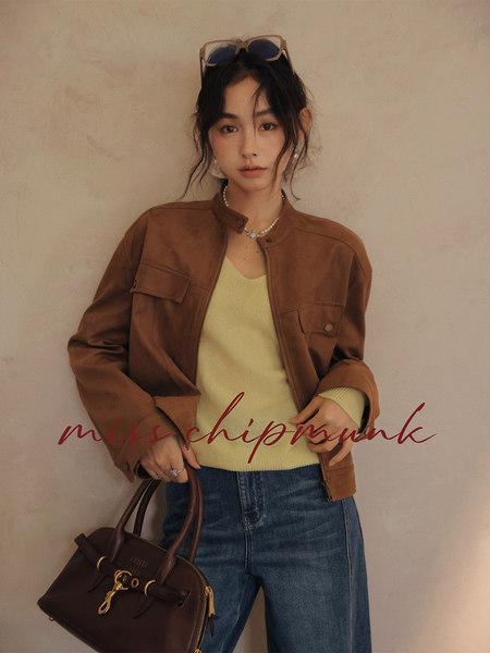 Woman wearing a brown jacket, yellow sweater, and blue jeans while holding a dark handbag