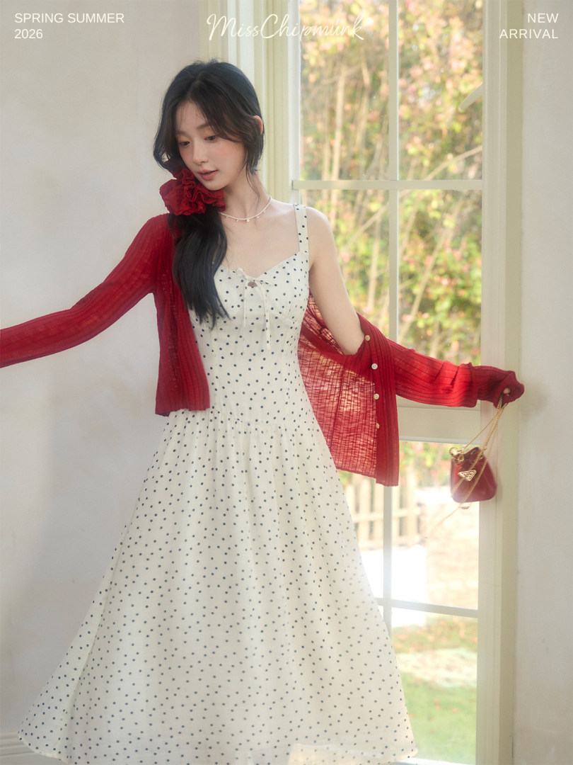 Model wearing a white polka dot sleeveless dress with a red cardigan by a window