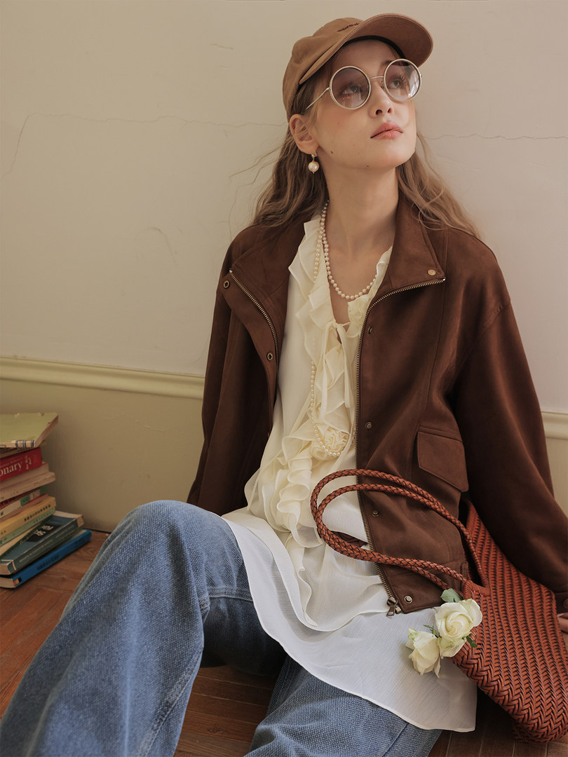 Woman wearing a brown jacket over a white ruffled blouse with jeans and a cap