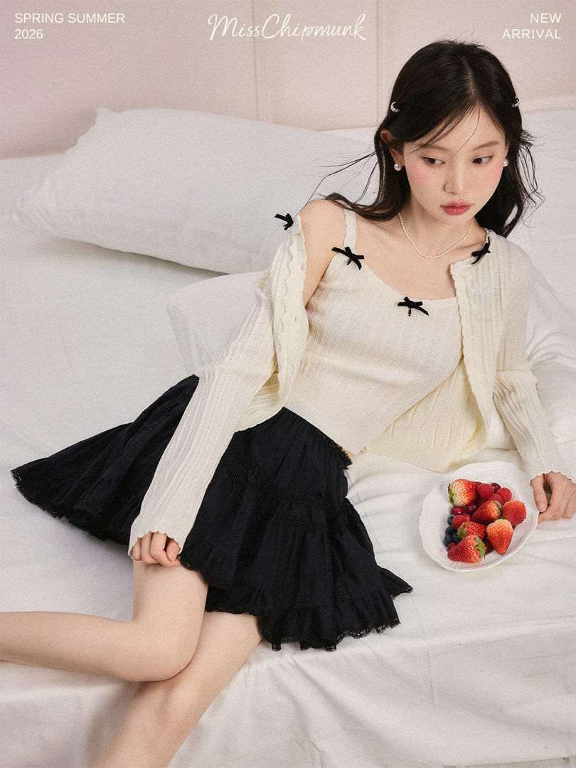 Model wearing a cream knit cardigan and top with a black ruffled skirt on a white bed
