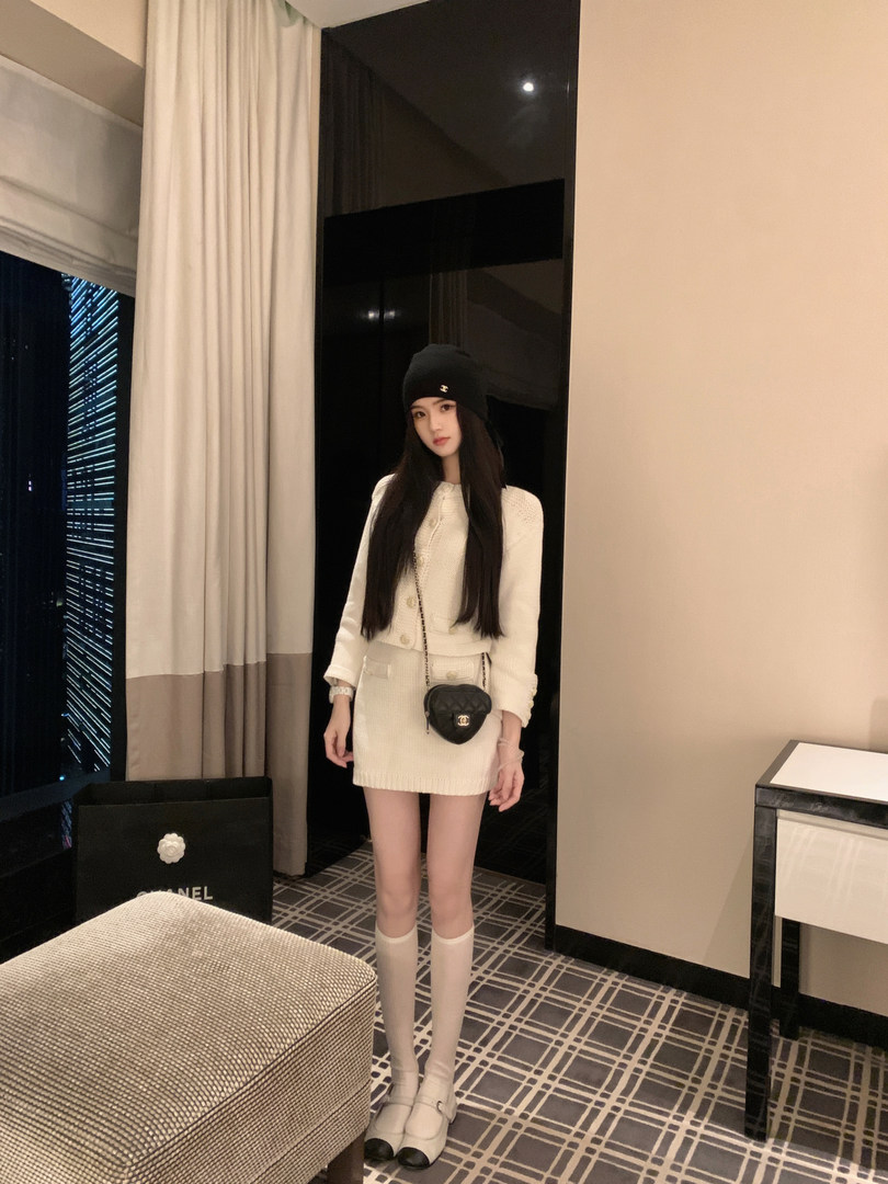 Woman wearing a white knit two-piece set with black beanie and heart-shaped bag, standing in a hotel room.