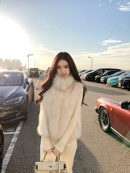 Woman wearing a white fur vest and matching outfit, holding a handbag, standing in a parking lot with cars and city skyline in the background.