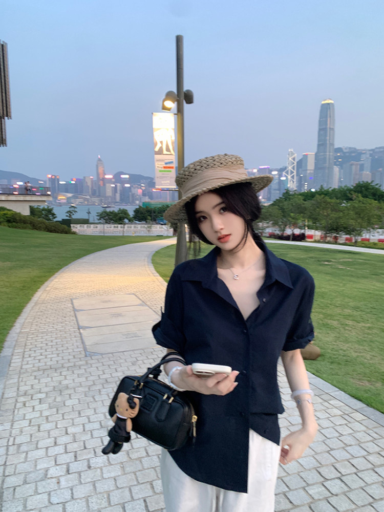 Woman wearing a navy button-up shirt, white pants, and straw hat, holding a black handbag with a city skyline in the background.