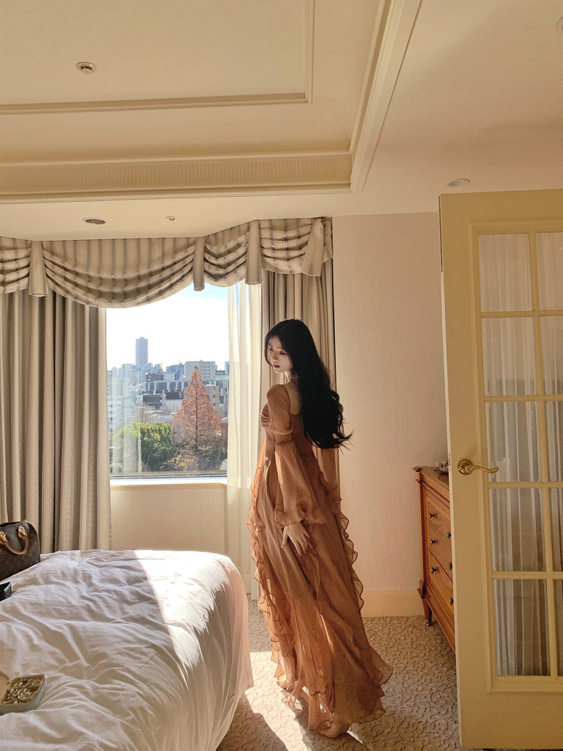 Woman wearing a long, flowing, ruffled dress in a hotel room with a city view.