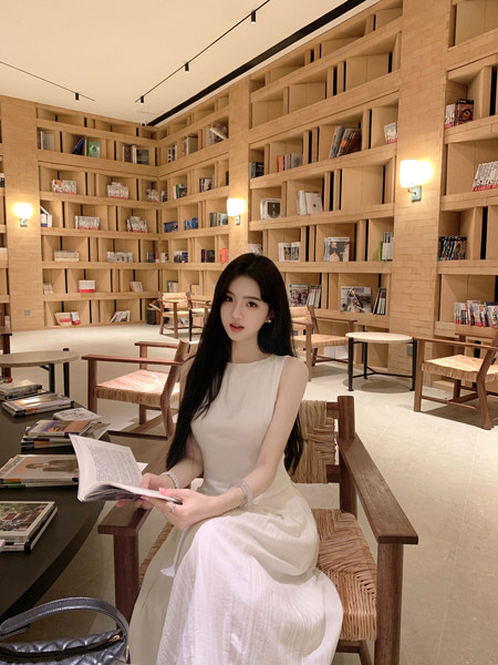 A woman in a white dress sitting in a library holding a book.