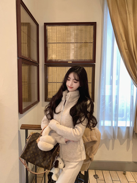 Woman wearing a cream-colored fuzzy zip-up jacket and matching pants, holding a brown monogram handbag with plush charms, seated indoors near framed plaques and a window.