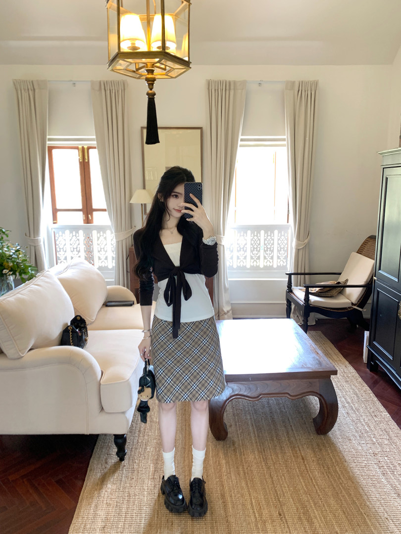 Woman wearing a black cardigan, white top, and plaid skirt in a living room.