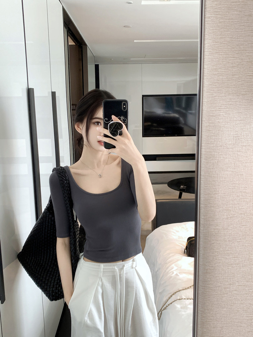 Woman wearing a dark gray fitted scoop neck top with white pants, taking a mirror selfie.