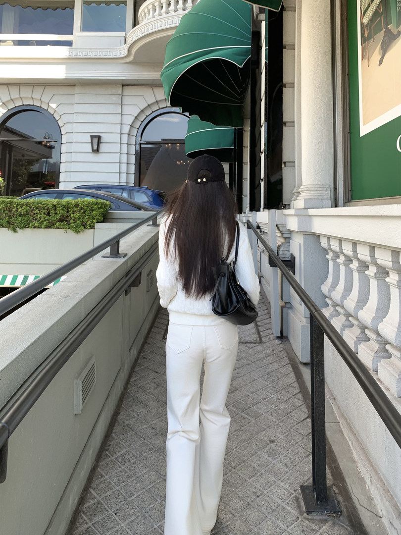 Woman wearing white pants and sweater with black bag walking on a sidewalk