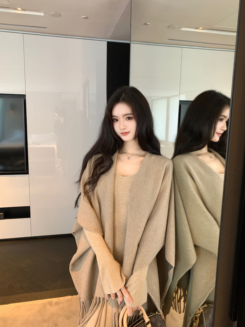 Woman wearing a beige knit poncho with fringes, paired with a ribbed top and holding a handbag, standing in front of a mirror.