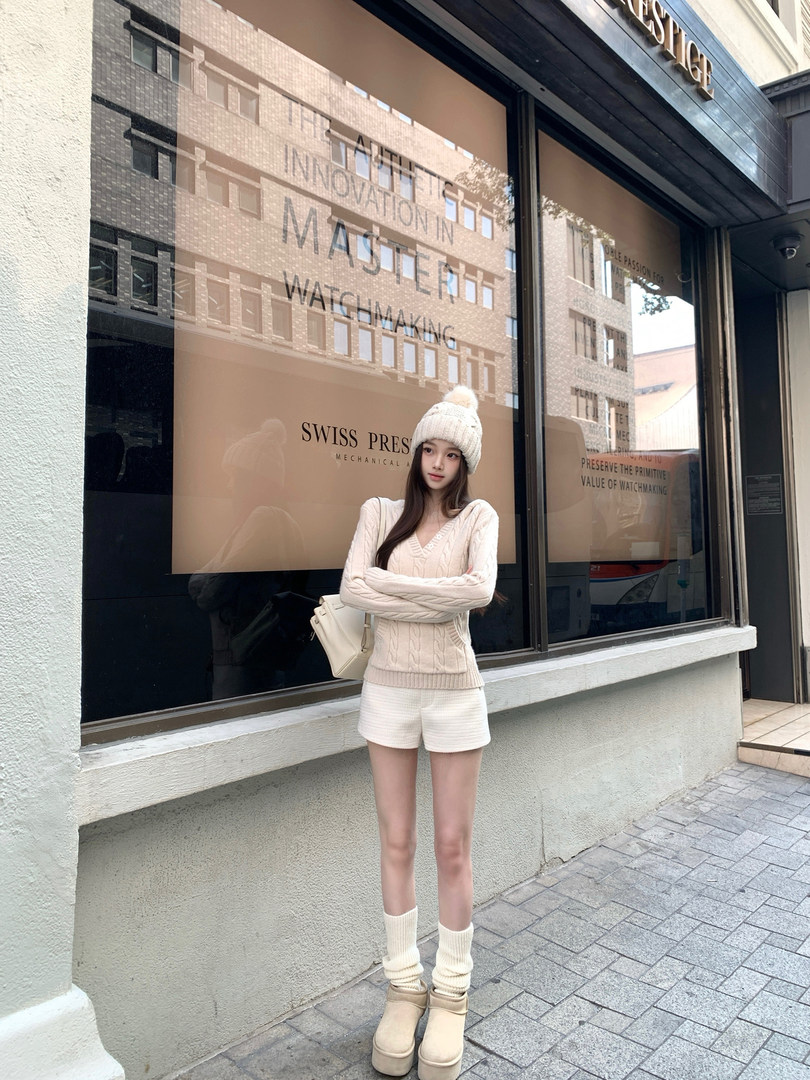 Model wearing cream cable-knit sweater, matching shorts, beanie, and boots standing outside a store.