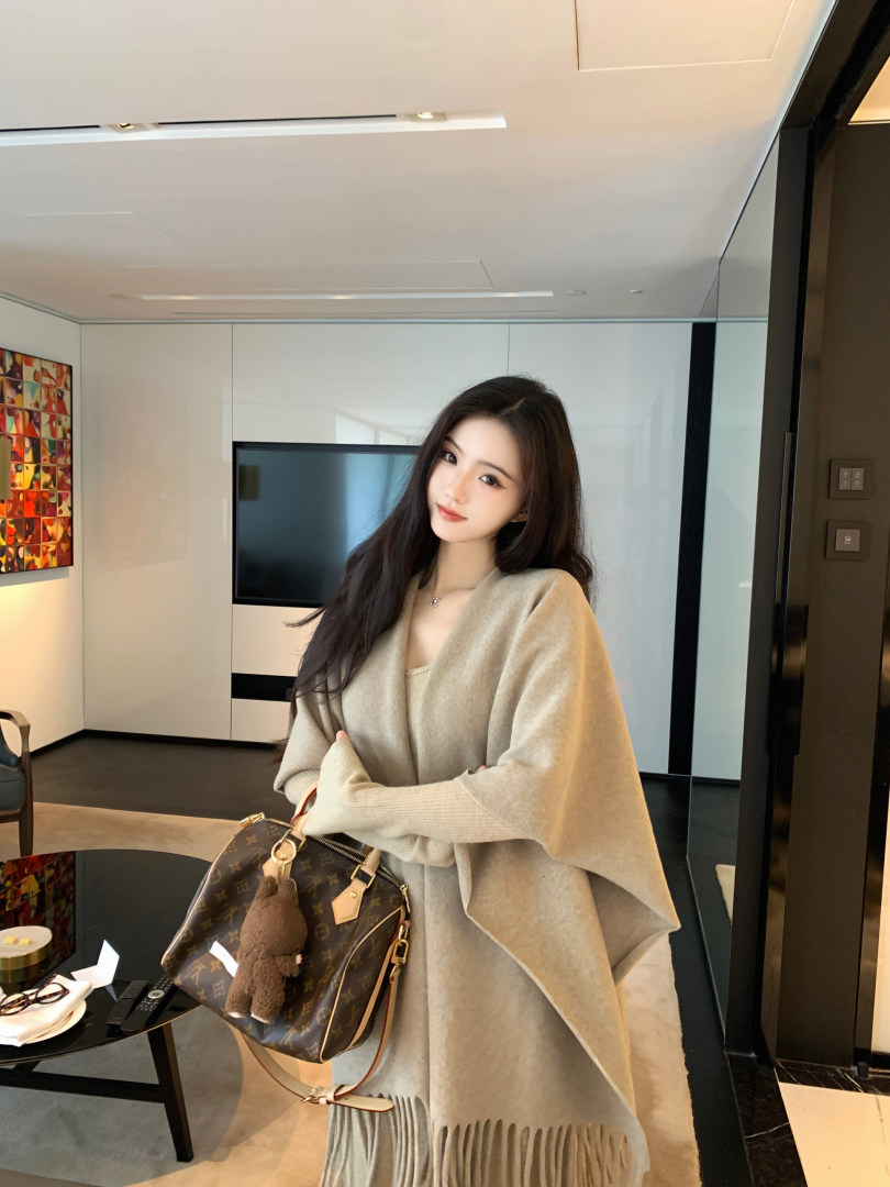 Woman wearing beige knit poncho with fringe, holding patterned handbag in modern interior