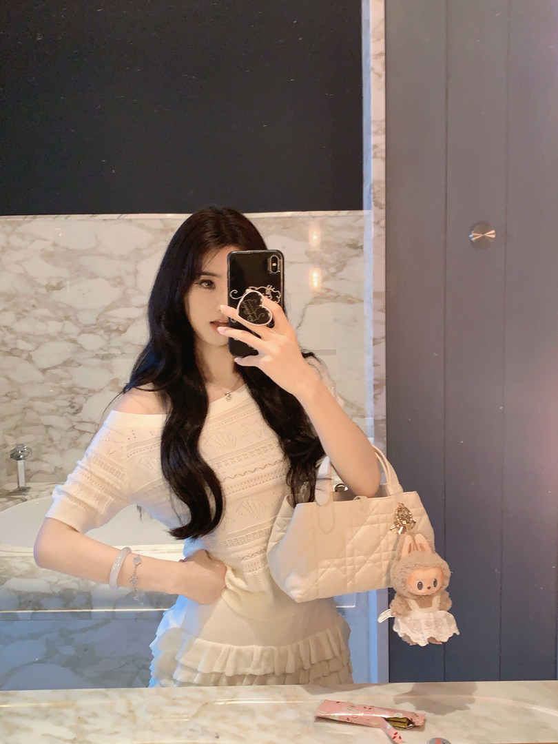 Woman taking mirror selfie wearing white off-shoulder top and holding a quilted handbag with a plush keychain.