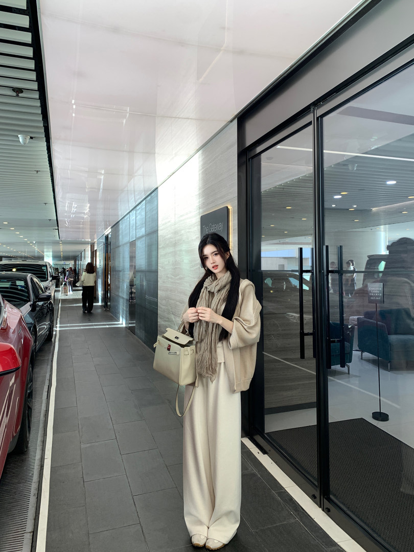 Woman wearing beige knit cardigan, scarf, wide-leg pants, and carrying cream handbag in modern building hallway