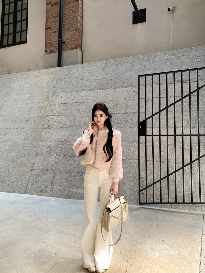 Woman wearing a pink faux fur jacket, cream wide-leg pants, and holding a beige handbag, standing against a concrete wall.