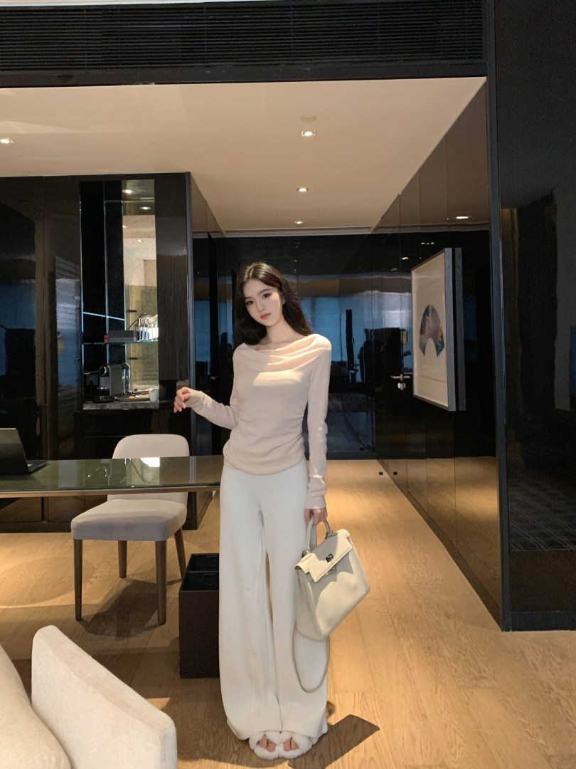 Woman wearing a beige long-sleeve top and white wide-leg pants, holding a white handbag in a modern interior.