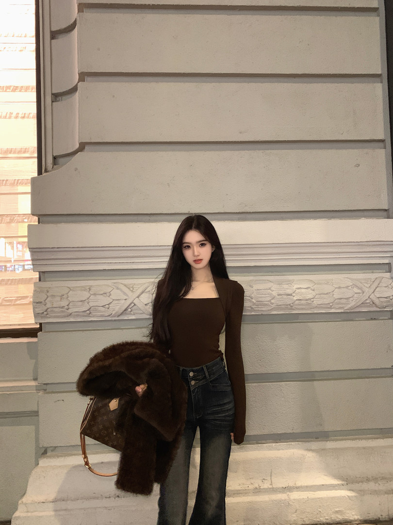 Woman wearing brown long-sleeve top and dark jeans, holding a fur coat and designer bag, standing against a building facade